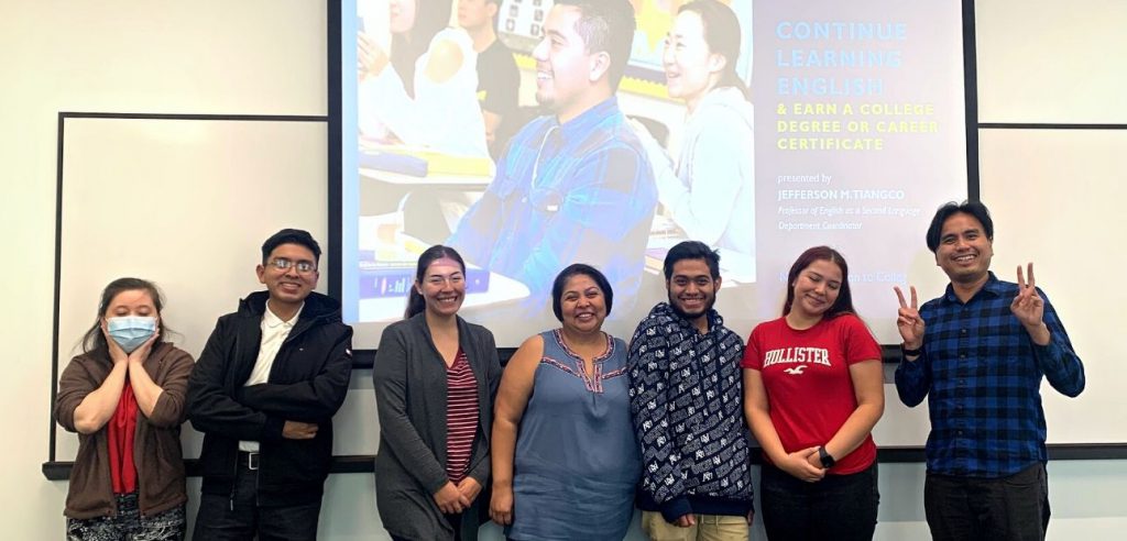 Siete estudiantes posan frente a una poderosa presentación en la pantalla del salón de clases que dice: "continúe aprendiendo inglés y obtenga un título universitario o un certificado profesional".