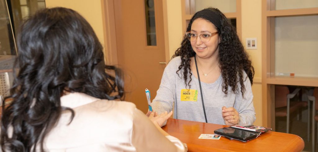 Una estudiante con anteojos y cabello rizado castaño oscuro que recibe asistencia para registrarse en el escritorio de Admisiones y Registros de Anaheim.