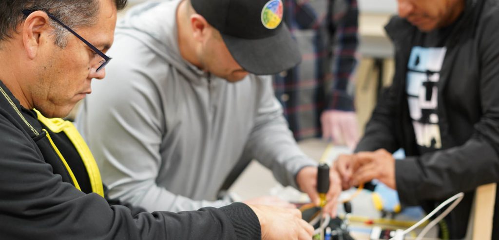 Una foto de dos estudiantes de electricidad. Uno asegura el interruptor de la luz y revisa los cables, mientras que el otro estudiante conecta el interruptor a la fuente de energía eléctrica principal.