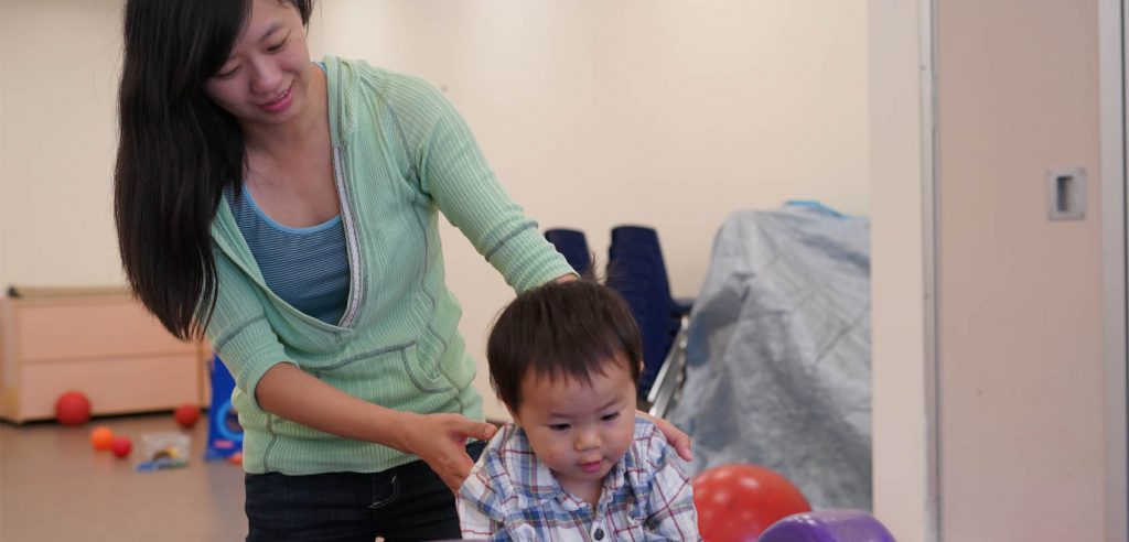 A photo of a mother lightly holding her toddler, making sure he doesn’t fall over. 