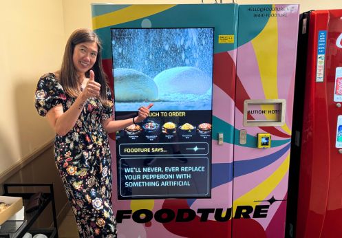 Student Leader, Lourdes Valiente is posing with two thumbs up in front of the Foodture vending machine. She is wearing a black and colorful flowery dress.