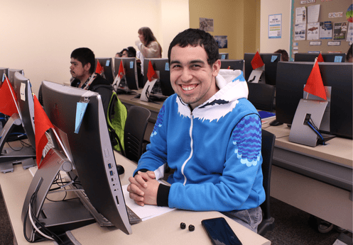 A smiling male DSS student sitting at a computer. He is of light complexion, dark short hair and wearing a blue and white yeti like hoodie.