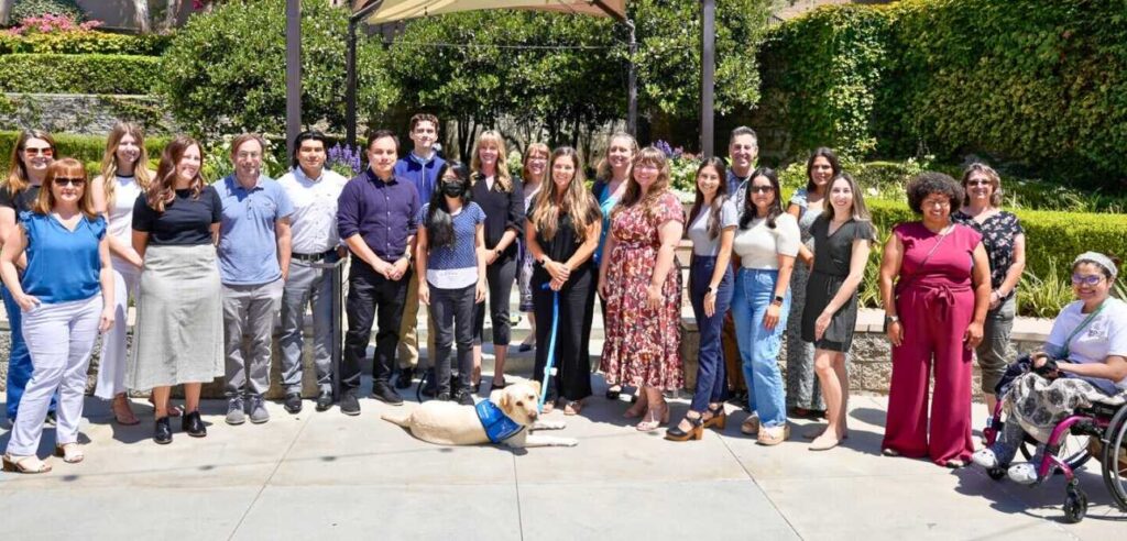 A group of 22 DSS staff members posing for a photo in front of green and pink bushes, with a facility dog lying in front of them.