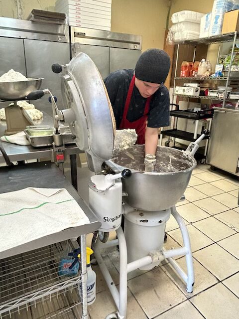 A student wearing a black shirt, red apron, and black beanie. This student is scooping out dough from a mixer.