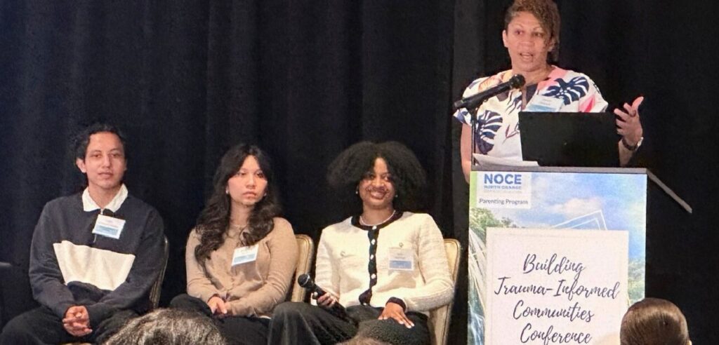 Three college students seated on stage listen as a standing speaker talks at a conference.