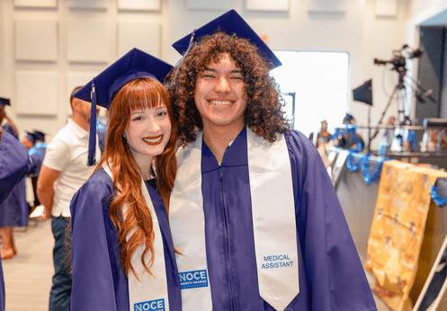 Two graduating Medical Assistant students posing together for a photo. One has long red hair, the other has brow curly hair. Both students are wearing dark blue cap and gowns with a white graduating sash.