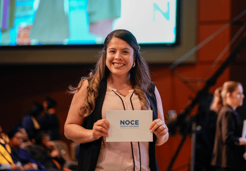 A English as a Second Language student posing for a photo with with her NOCE graduation card. She has brown curled hair and is wearing a black vest, and a light pink blouse.