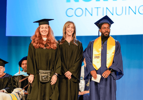 A high school diploma student posing for a photo with two of his teachers on stage. The student is wearing dark blue cap and gown with a gold graduating sash.