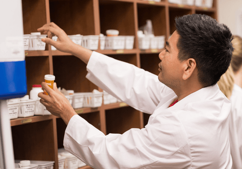 A Pharmacy Technician student with short dark hair placing extra medications in bins that are located on a shelf.