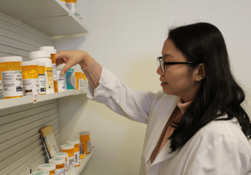 A Pharmacy Technician student with long dark hair and glasses checking labels on medications that are located on a shelf.