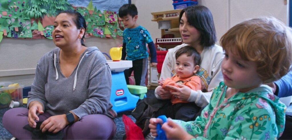 Two mothers sit cross-legged on the floor with three young children in an early childhood classroom looking with interest in a relaxed manner toward the teacher (not shown).