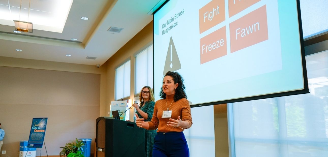 Jamie De La Mora is speaking at the 2026 Building Trauma-Informed Communities Conference, with a slide showing stress responses: Fight, Freeze, Fawn. With Dr. Erin Sherard standing behind her at a podium.