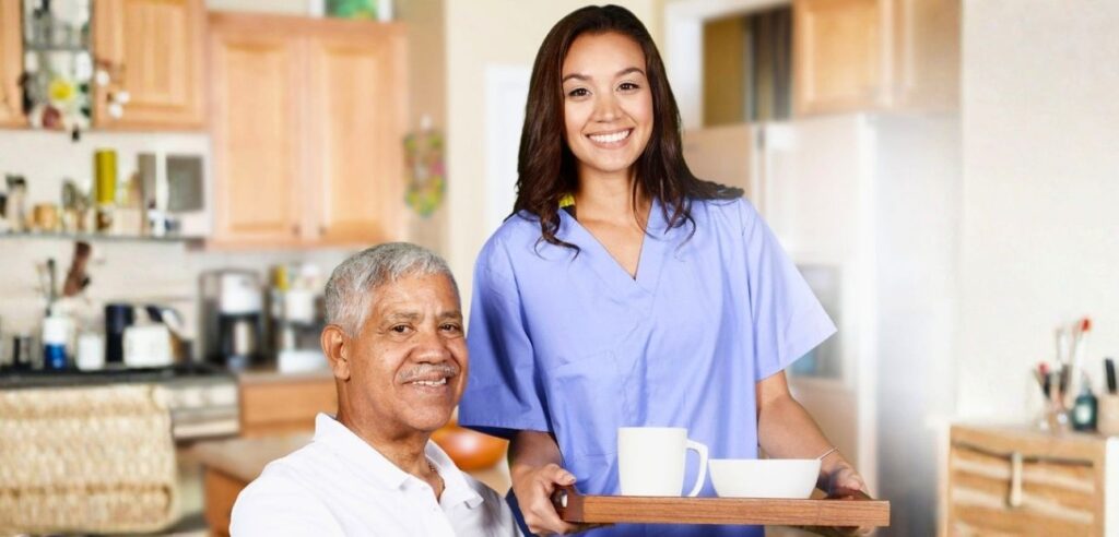 Young female personal care aide in blue scrubs serving coffee to elderly man at home, demonstrating caregiving skills.