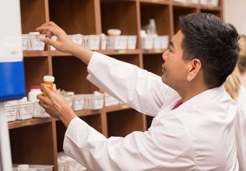 Pharmacy technician selecting medication bottle from organized shelving unit in clinical pharmacy setting