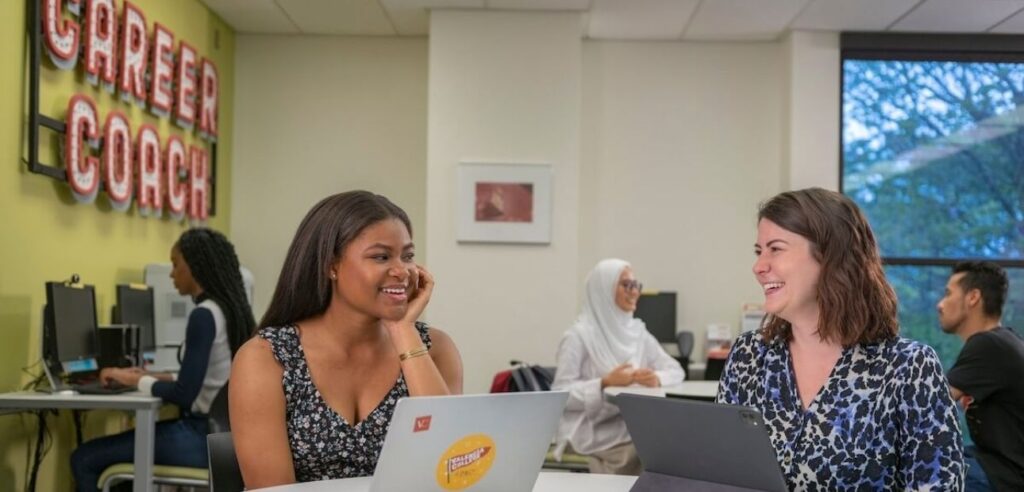 Two smiling female students on their laptop and tablet, having a fun conversation.
