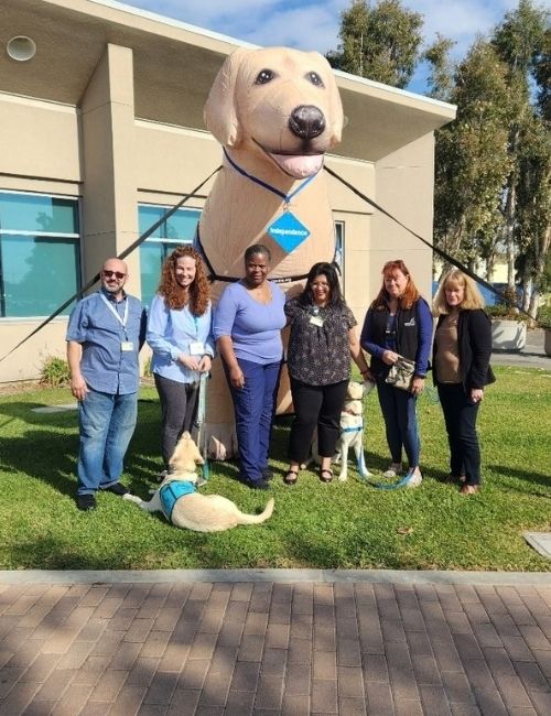 A large inflatable dog mascot wearing a blue collar stands on a lawn in front of a building, while six people pose for a photo, with trees and a blue sky in the background.