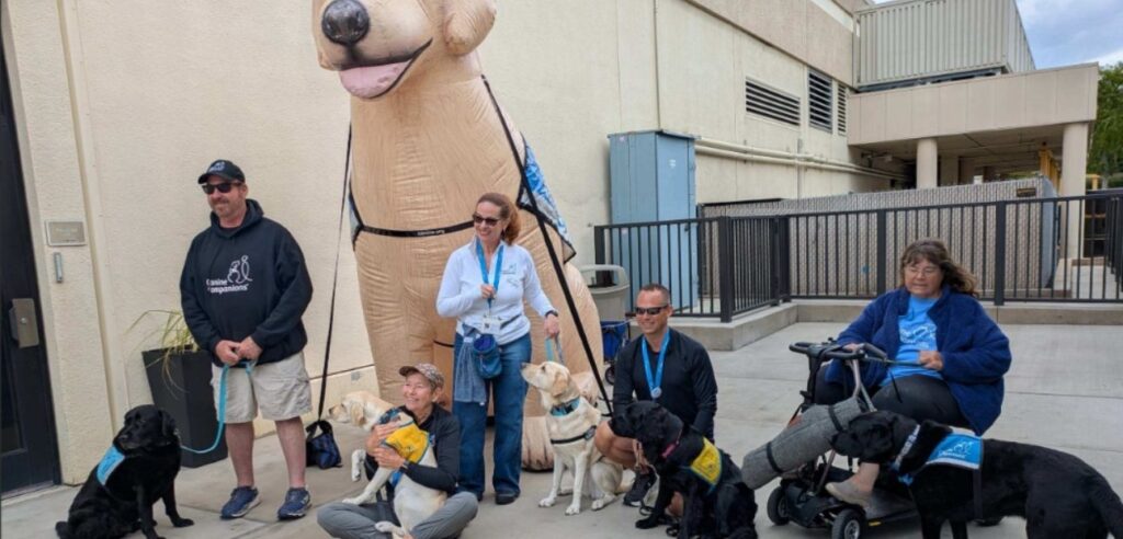 A group of people with several service dogs pose outdoors in front of a large inflatable dog, with some handlers kneeling and others standing.