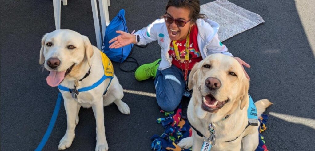 A woman in a red colorful shirt and glasses kneels on the pavement with two golden labrador retrievers wearing harnesses.