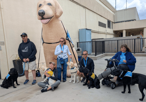 A group of people with several service dogs pose outdoors in front of a large inflatable dog, with some handlers kneeling and others standing.